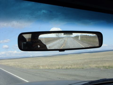 1988 Ford Bronco - View through the interior rearview mirror shows a long, straight road with dry grass fields on either side under a cloudy sky.