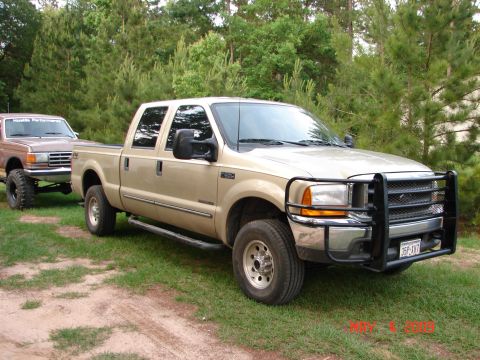 1987 Ford Bronco - Two pickup trucks, a gold Ford F-250 and a brown lifted truck, are parked on a grassy area with trees in the background.