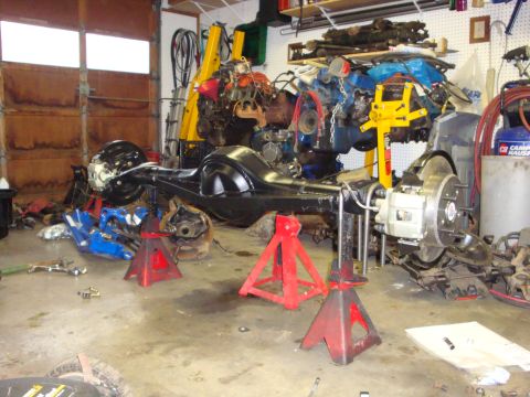 1989 Ford Bronco - Rear axle assembly, likely from a 1989 Ford Bronco, is supported by jack stands in a garage setting, with an engine and hoist nearby.