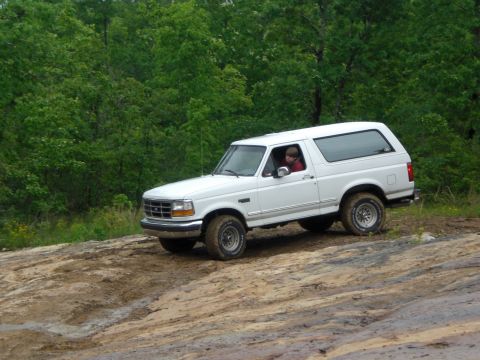 1989 Ford Bronco - A white 1992 Ford Bronco navigates a muddy, off-road trail with trees in the background.