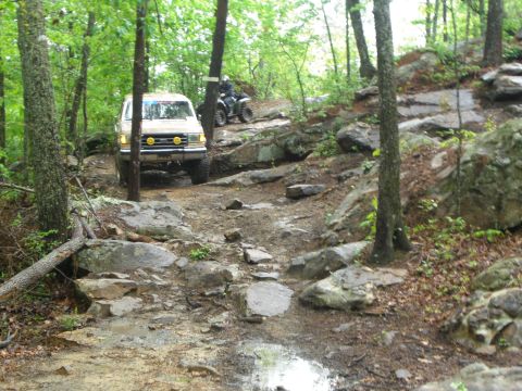 1989 Ford Bronco - A tan Ford Bronco tackles a challenging off-road course, with an ATV visible further up the path.
