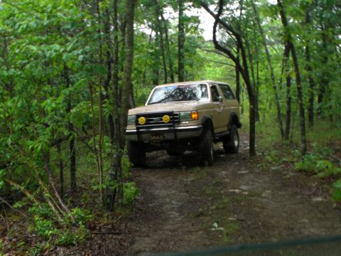 1989 Ford Bronco - A lifted Ford Bronco navigates a wooded trail, kicking up mud on the path ahead.
