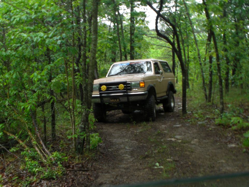 Tan Ford Bronco with off-road tires driving on a muddy trail through a forest. It has yellow auxiliary lights on the front