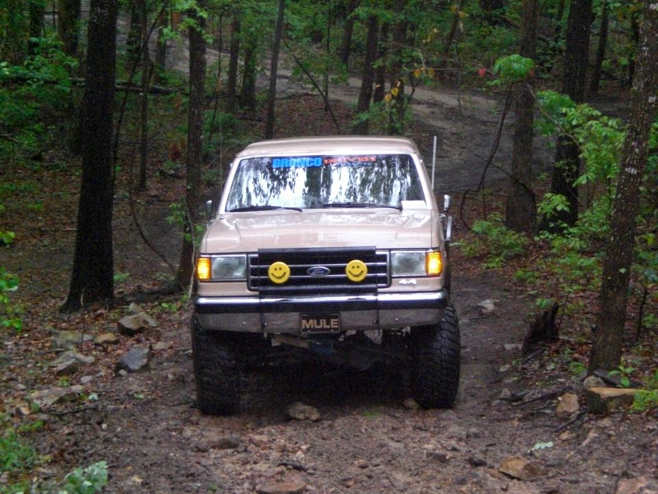 Tan Ford Bronco with smiley face headlights and "MULE" license plate driving on a dirt path in a forest