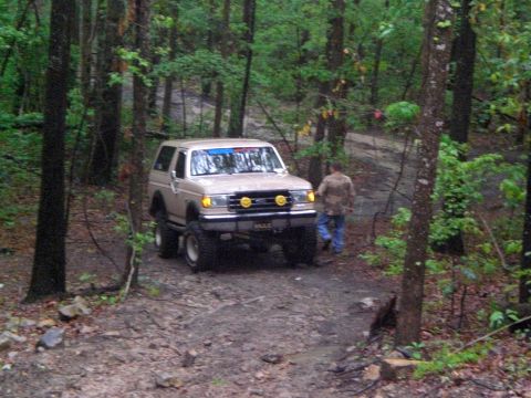 1989 Ford Bronco - A tan 1989 Ford Bronco navigates a muddy, wooded trail with a person walking nearby.