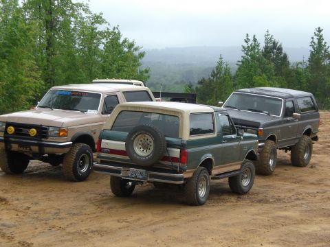 1989 Ford Bronco - Three Ford Bronco SUVs are parked on a dirt road, with a wooded landscape visible in the distance.