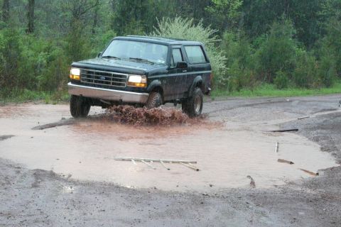 1995 Ford Bronco - A black SUV navigates a large muddy puddle, kicking up water and dirt.