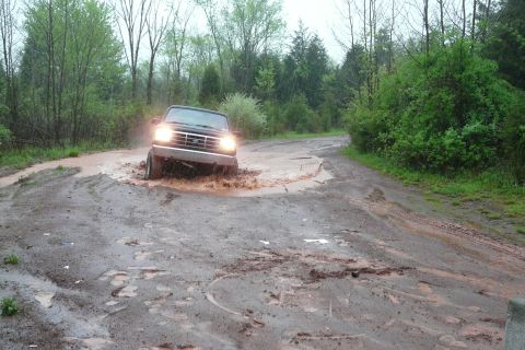 1995 Ford Bronco - A dark-colored Ford Bronco navigates a water-filled rut on a wet, wooded road.