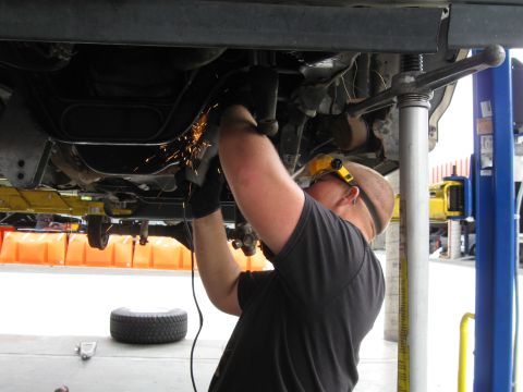 1986 Ford Bronco - A mechanic is working on the underside of a vehicle, cutting metal with an angle grinder.
