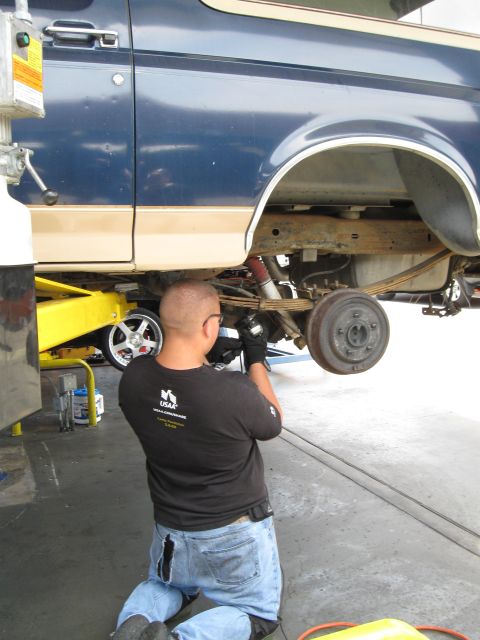 1986 Ford Bronco - Mechanic performs work on the rear suspension of a blue and tan SUV, possibly from the 1980s.