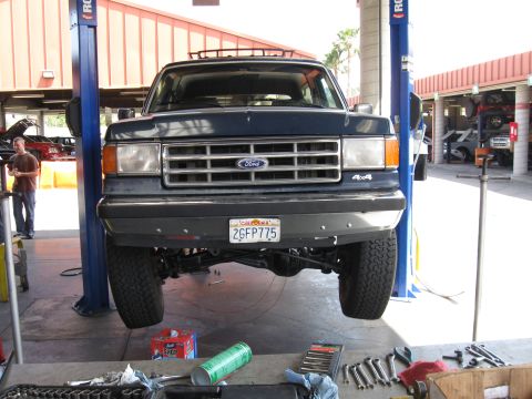 1986 Ford Bronco - A dark blue 1987 Ford Bronco is raised on a hydraulic lift in a repair shop, showing its undercarriage and front grille.