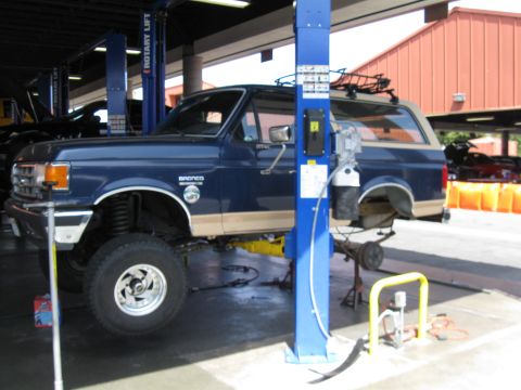 1986 Ford Bronco - A blue 1986 Ford Bronco with tan trim is shown on a lift in a service bay, undergoing maintenance.