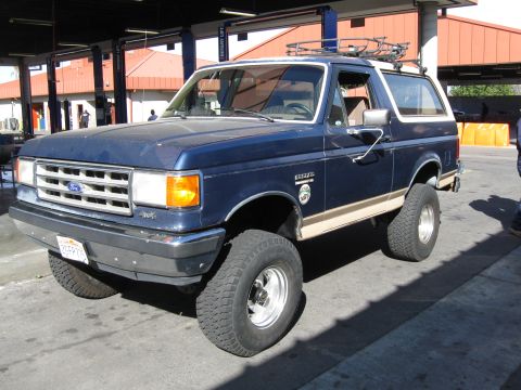 1986 Ford Bronco - A lifted blue Ford Bronco with a tan stripe is parked outside a building with car lifts.