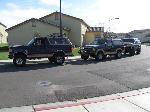 1986 Ford Bronco - Two lifted Ford Bronco SUVs are parked side-by-side on a suburban street.