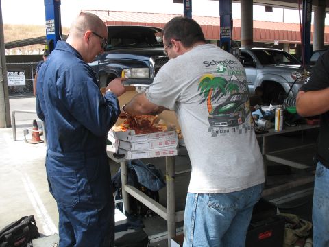1986 Ford Bronco - Men share pizza in a garage setting, with vehicles present in the background.
