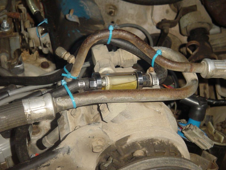 Close-up of a clear inline fuel filter with yellow fluid, connected by hoses and zip ties in an engine bay