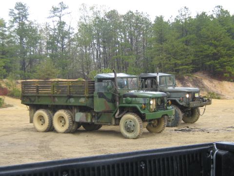 1990 Ford Bronco - Two military trucks are parked on a dirt lot with trees in the background.
