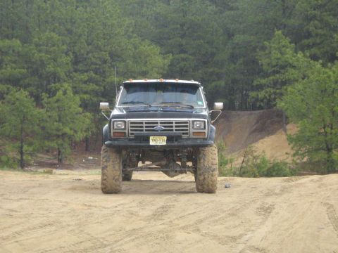 1990 Ford Bronco - This lifted Ford pickup truck is shown from the front, parked on a dirt road with trees in the background.