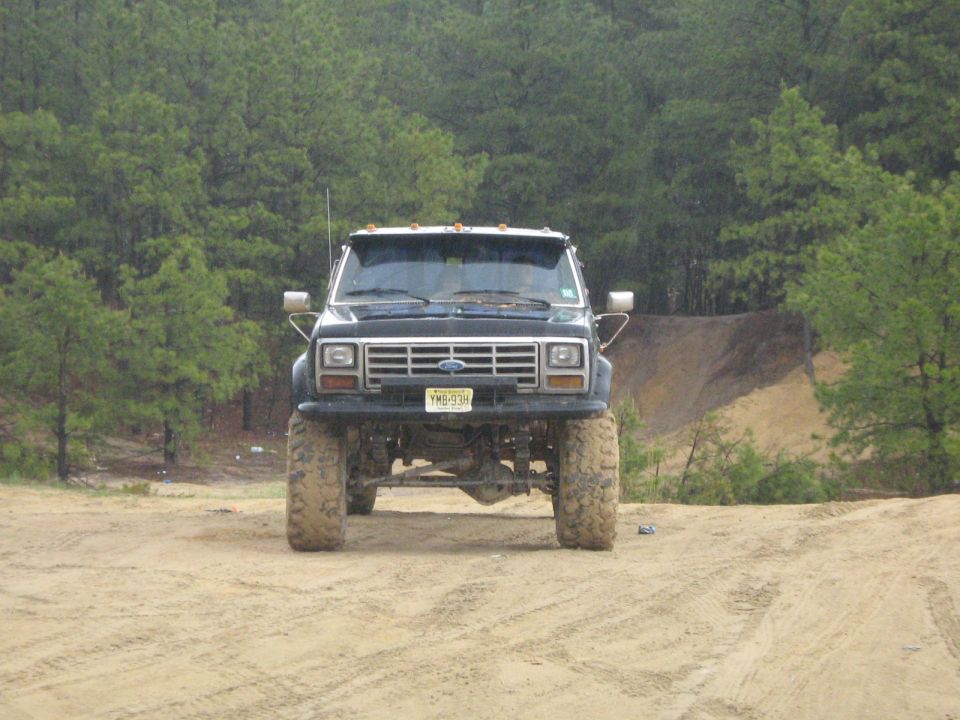 Front view of a lifted black Ford pickup truck with large tires on a dirt road, trees in background