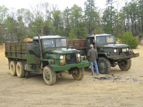 1990 Ford Bronco - Two military trucks are parked on a dirt lot with trees in the background.