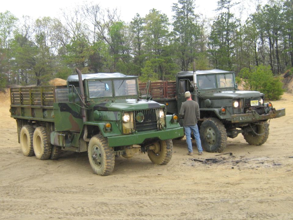 two green military trucks parked on dirt, one with a man standing between them