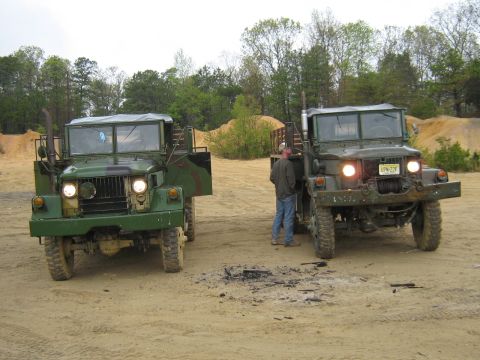 1990 Ford Bronco - Two military trucks are parked on a dirt lot with trees and sand piles in the background.