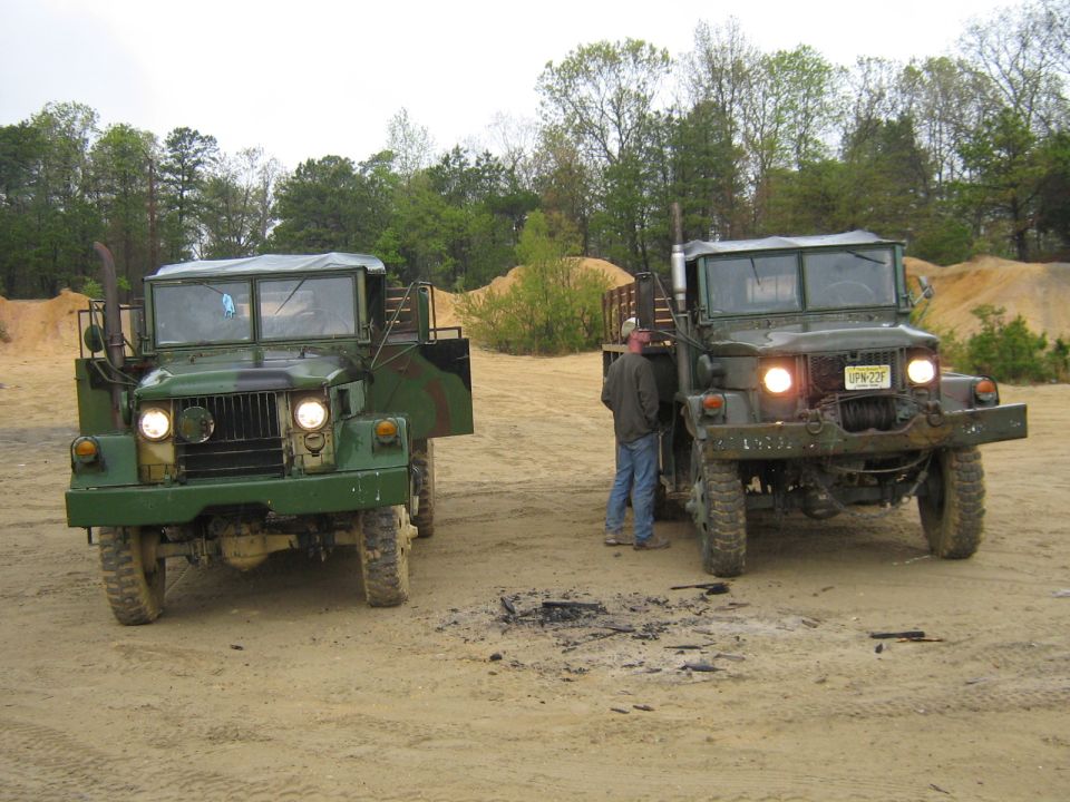 Two green military trucks parked on dirt, one with its door open. A man stands between them
