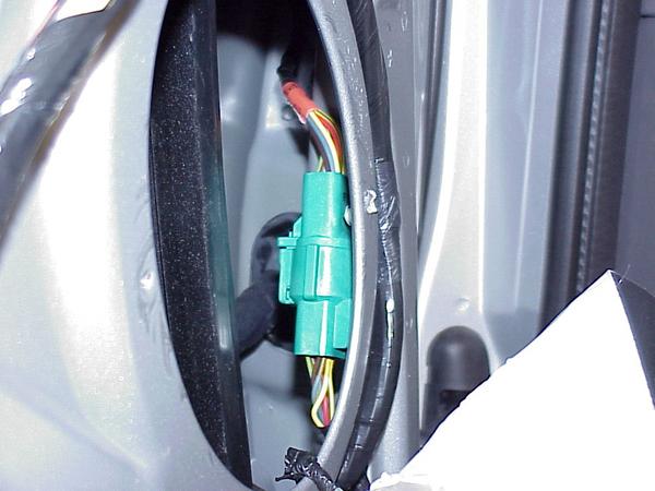 Close-up of a green wiring harness connector with multi-colored wires, attached to a gray vehicle interior