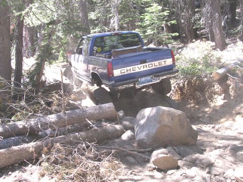 1994 Ford Bronco - Chevrolet pickup truck is off-roading on a rough trail with fallen trees and large rocks.