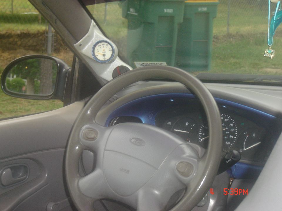 Gray steering wheel with Ford logo, dashboard gauges, and aftermarket gauge pod. Exterior view of trash cans and fence visible through windshield