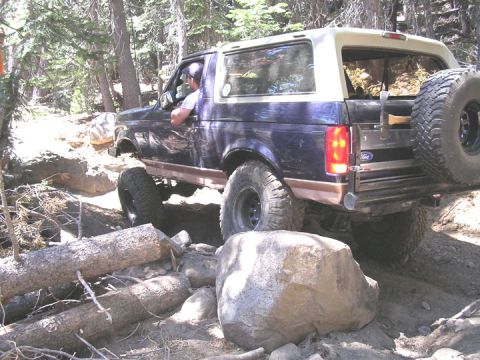 1994 Ford Bronco - A dark blue Ford Bronco with a tan top navigates a challenging, rocky off-road trail through a wooded area.