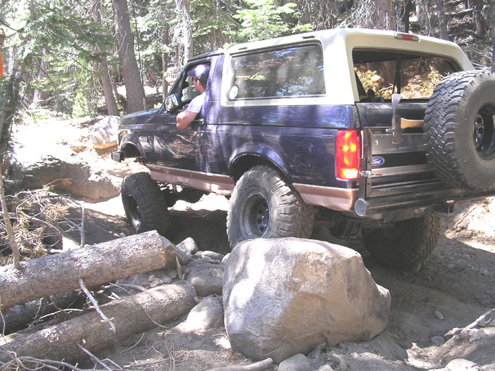 Blue Ford Bronco SUV with tan top crawling over rocks and logs on a forest trail