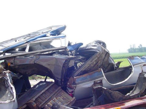 1992 Ford Bronco - Severely crushed and rolled automobile body resting in a salvage yard with farm fields in the background.