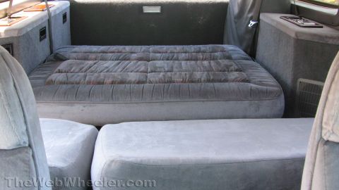 1996 Ford C-350 (Centurion Conversions) - Rear interior of a conversion SUV shot from between the front seats, showing a gray cloth third-row bench folded flat into a bed with the s…