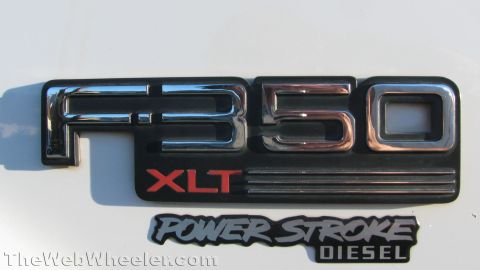 1996 Ford C-350 (Centurion Conversions) - Tight close-up of the chrome-and-black "F-350" fender badge with red "XLT" lettering on a white Ford truck panel, with a black "Power Strok…