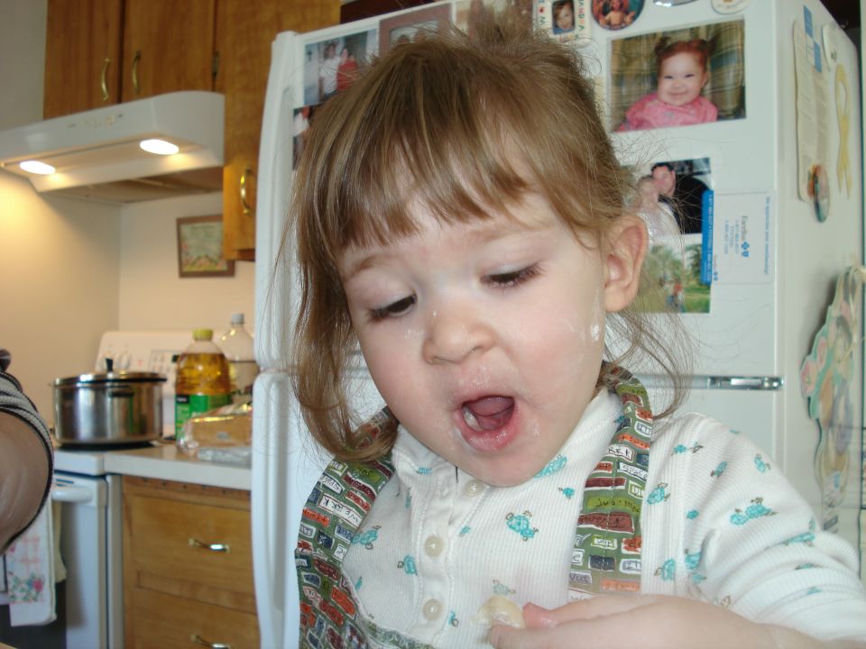 Toddler with flour on face, looking down with mouth open, in a kitchen with a refrigerator covered in photos