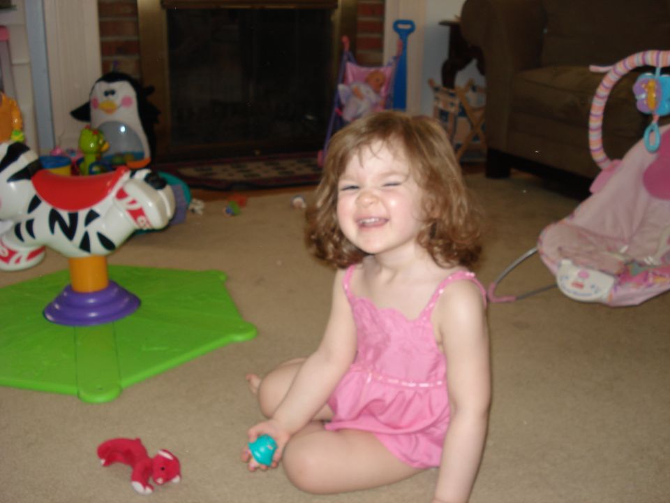 Child in pink dress sitting on carpet, holding a blue ball, with toys around