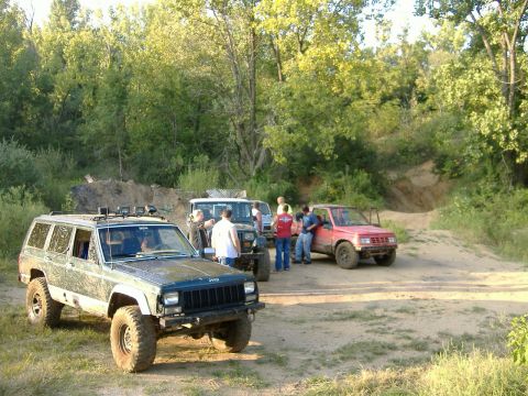 1993 Jeep Cherokee - Several off-road vehicles, including a 1993 Jeep Cherokee, are parked in a dirt area with trees in the background.
