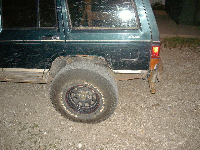 Dark green Jeep Cherokee rear quarter with BFGoodrich All-Terrain tire on black steel wheel, parked on gravel