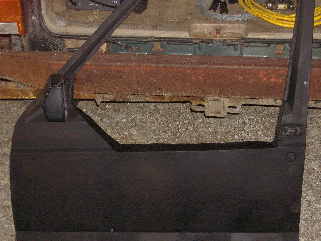 Close-up of a black Jeep Cherokee door panel, showing the interior side and part of the frame. The vehicle's rusty rear bumper is visible behind it