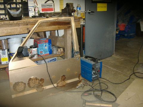 1987 Jeep Comanche - A heavily rusted Jeep door panel sits on a workbench next to a welding machine in a garage.