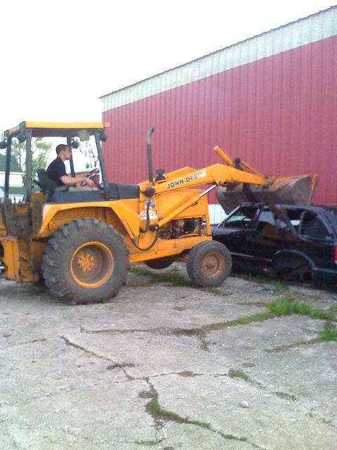 1992 Ford Bronco - A yellow John Deere backhoe loader is positioned next to a black SUV in front of a red building.