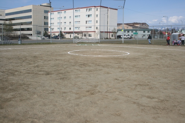 dirt softball field with bases marked, buildings in background