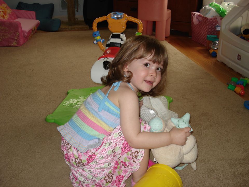 Child in a colorful dress kneels on a carpeted floor, holding a stuffed animal. Toys are scattered around