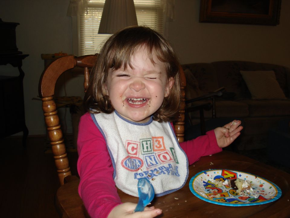 Child with messy face and bib, holding a spoon, with a plate of cake