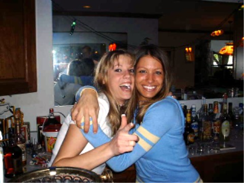 1990 Ford Taurus - Two women embrace at a bar, with a reflection in the mirror behind them and bottles lining the counter.