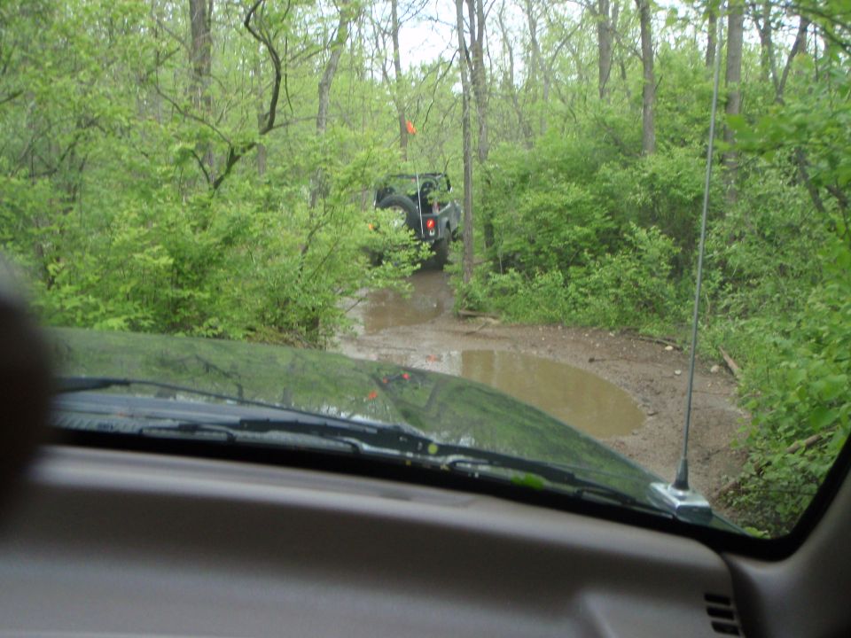 View over black hood and tan dash of a muddy wooded trail with a 4x4 ahead
