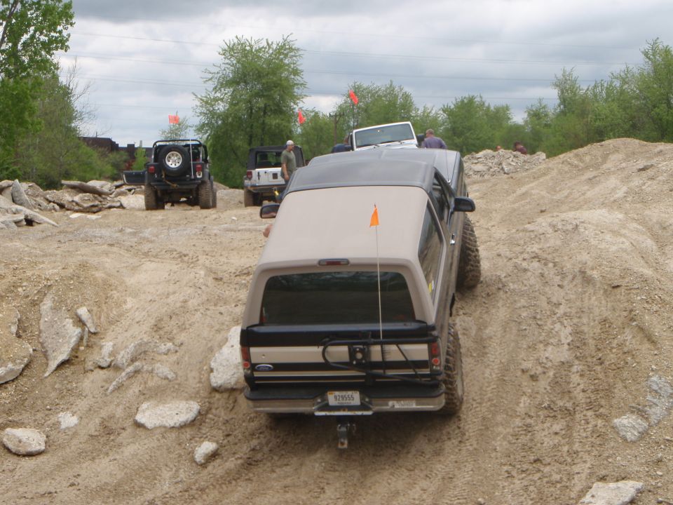 Tan and black Ford Bronco climbing a rutted dirt trail with Jeeps in background