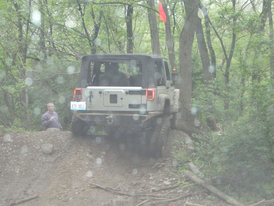Silver four-door Jeep Wrangler with soft top climbing a rutted wooded dirt trail