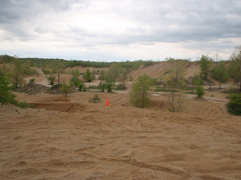 Sandy terrain with tire tracks, trees, and hills under a cloudy sky. An orange cone is visible